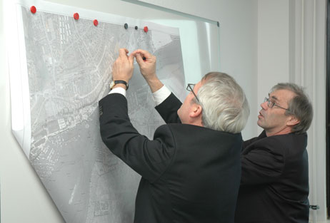 Wolfgang Wiese (Lübeck Port Authority) und Michael Lange (Stabsstelle Sonderaufgaben und Beteiligungen) bringen für die Planungen ein Luftbild der Trave an der großen Magnettafel an. Foto: TA